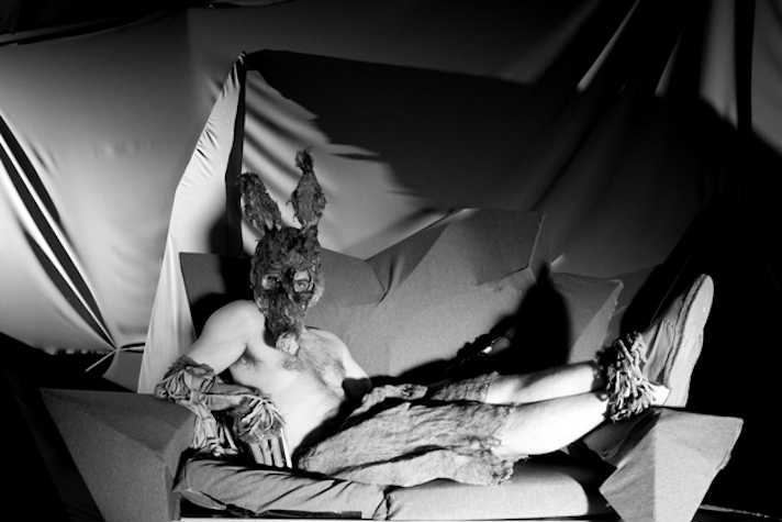 black and white film still of a man laying on a geometric, sculptural sofa, topless but wearing shorts. he also wears a mask that resembles a rat or rabbit and looks towards the camera.