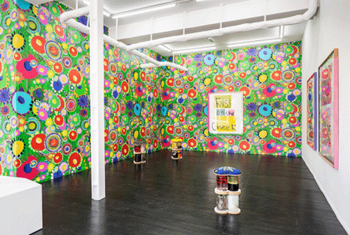 The image shows a colourful exhibition with bright patterned wallpaper and sculptures made of glass jars