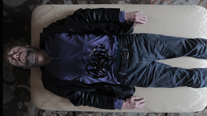 A man is lying down on a settee, with his eyes closed. There is a tubular sculpture laid on his stomach.