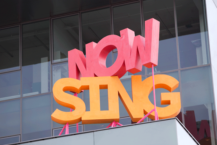 The words "Now Sing" are standing on stands, as a bold block sculpture. They are bright coloured. They are set on the terrace of a modern building.