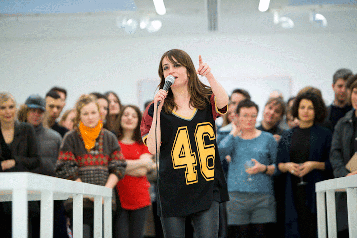 A woman is standing in the middle of the room with a full crowd behind her. She is speaking into a microphone and pointing to and looking at somewhere in the distance. There are long tables placed on either side of the woman.