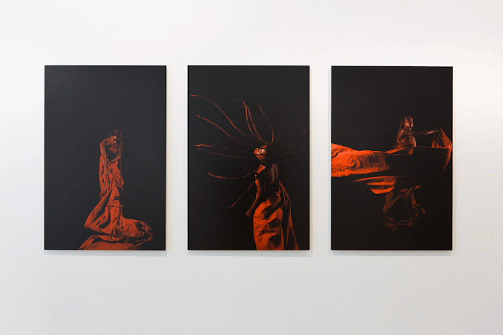 three photographs of a black person making dynamic movement swathed in red fabric. In the centre image braids are suspended in the air as they throw their head, in another fabric is suspended across the image as they pull it through the air