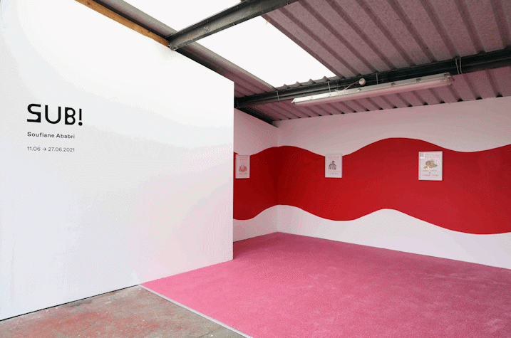 a wide red squiggle painted on a gallery wall. Small framed drawings hung on the wall. A pink carpet and pile of books