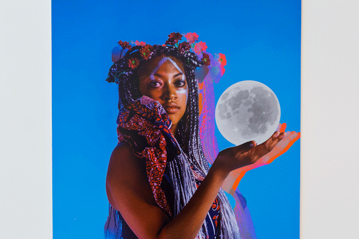 portrait photograph of a person of colour on bright blue background with flowers in their hair and moon in their hand