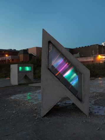 geometric concrete structure with different coloured lights streaming across the centre. it is situated on an empty outdoor lot with low buildings in the background