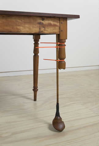 close up of a wooden table. one of the legs has been snapped off halfway and replaced with a wooden golf club, secured by red elastic bands,