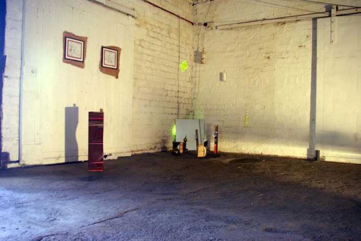 warhouse type space with two small framed works hanging on one wall. there is plinth beneath them, and a mirror placed on the floor in the corner with objects in front of it.