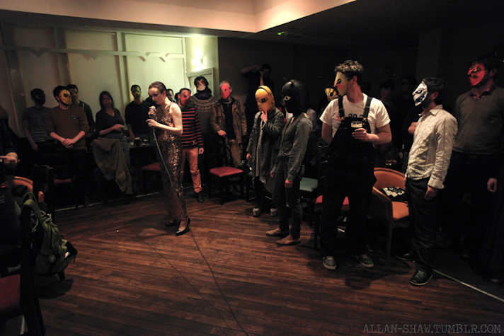 performance by Michelle Hannah. One person is in the centre of the stage holding a cabled microphone. everyone else in the room wears wrestling masks.