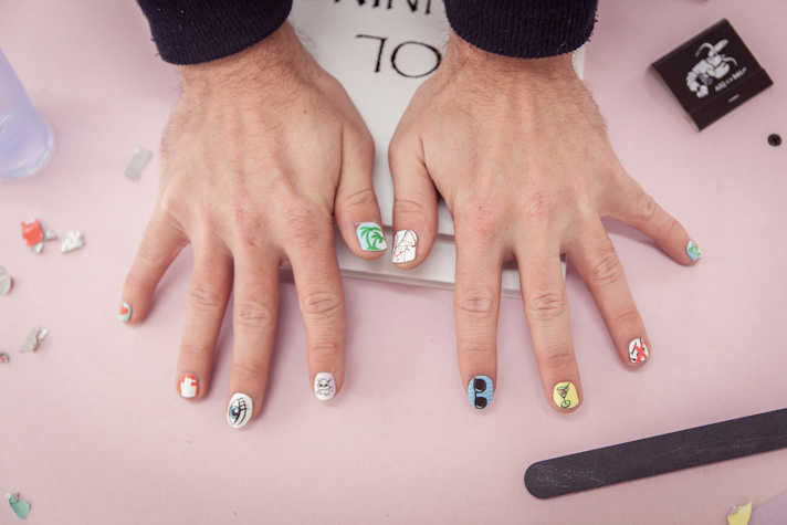 On a bright table, a mans's hands are spread out showcasing nail art done on their nails. Each nail has a different pattern; one has a pair of glasses made on it, the other had an eye, while another has trees. There is a nail file in the low right corner, a booklet under the hands and nail paint remover on the left.