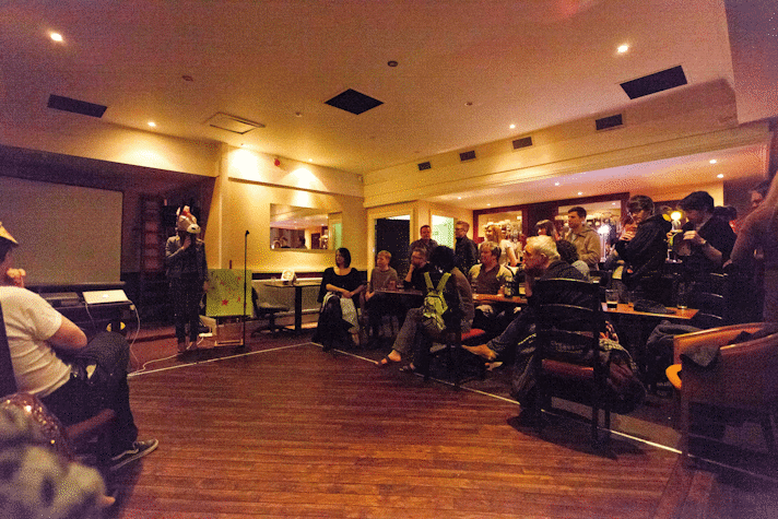 photograph of performance. the performer wears a horse mask as the audience watch on straddled on both sides of the makeshift stage