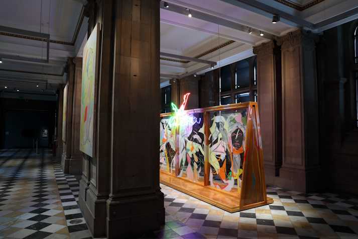 An abstract neon light showing two legs kicking the air in pointed heels, hangs at the top of a large wooden structure with glass panels and covered in fast gestural line paintings of figures and faces