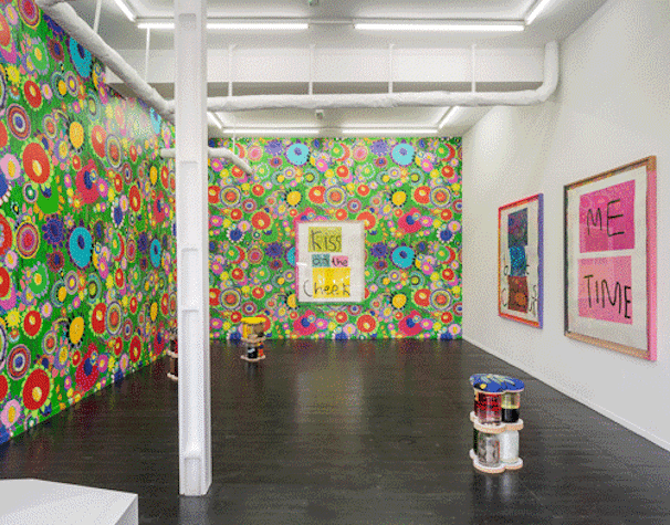 The image shows a colourful exhibition with bright patterned wallpaper and sculptures made of glass jars