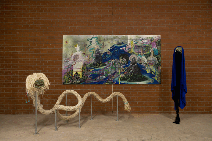 There is a rectangular, landscape-oriented painting on paper, hanging on a red brick wall. There are surreal, psychedelic faces merging with and emerging out of a landscape. There is the edge of an inky lake below a mountain, a crying face whose tears lead a river into another face's mouth. Next to the painting a mask with half closed eyes and a wide mouth is adorned with a cape of blue fabric, dark brown hair falls from its mouth. In front of the painting another mask has a rope body.