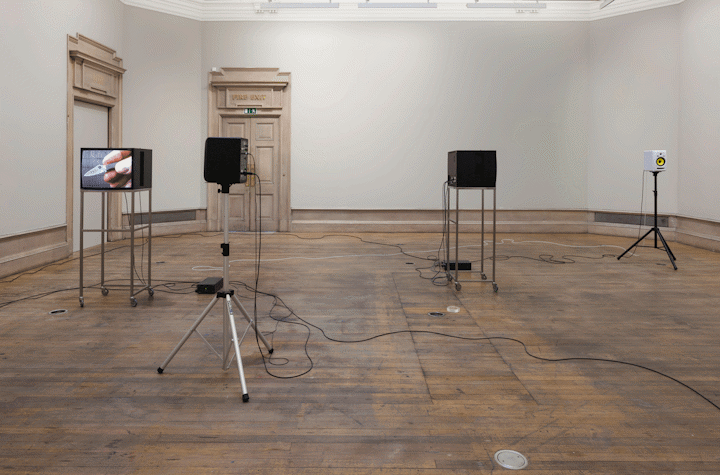 In a room with a wooden floor, there are two trolley stands with monitors and two tripods with speakers on them. These stands have wired connected to two control boxes in the middle of the room, and some following to the front tight corner of the image. One of the monitors has the image of fingers holding a small blade.