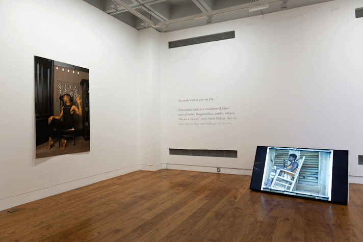 a large photographic print of a person in a dynamic pose. Black cursive text on an wall. A TV screen propped on the gallery floor.