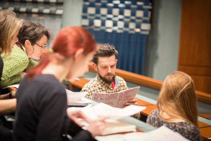 People are sitting on stationary benches in a lecture theatre. They are talking to each other while referring to the Glasgow International programme booklet.