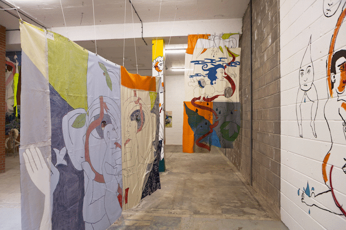 Playful and surreal line drawings of figures with blocks of colour cover a wall, column and fabric hanging in an industrial space