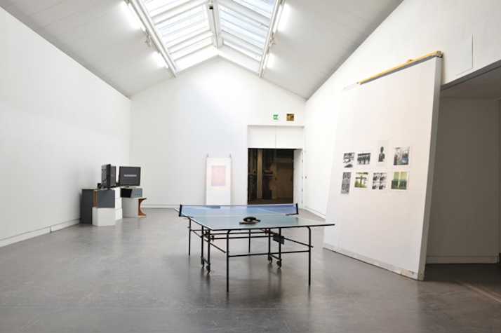 large white room with ping pong table in the centre. there are three TV monitors resting atop white boxes in the back left. on the right there is a display with 8 photographs.