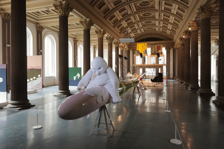 A gallery with a high vaulted ceiling and columns. In the middle a large, white toy-like cartoon figure sits on what appears to be rocket (sculpture). Other art works are visible in the background
