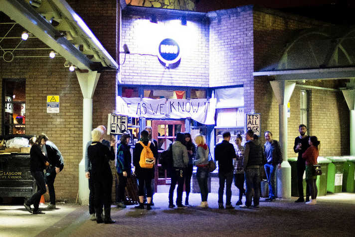exterior of the pipe factory at night with a small crowd outside. there is a banner which reads 'LIFE AS WE KNOW IT' and two signs reading 'THE END' and 'IT'S ALL OVER'