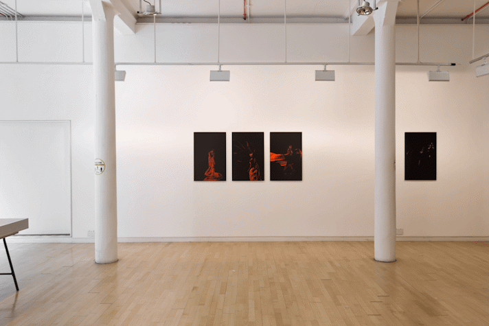 at a distance across a gallery a series of portrait photographs of a black figure on black background with red drape of fabric, hang on a wall