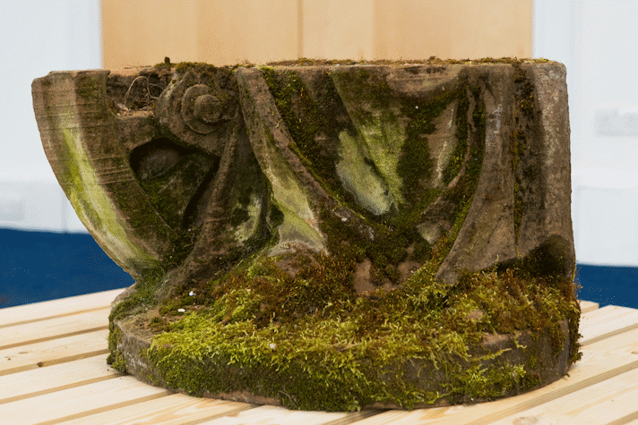 a section of a red sandstone sculpture covered in moss sits on a wooden structure