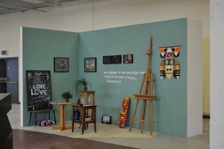 turquoise painted wall creating an interior scene. there is a easel, and picture frames leaning against the wall, with more frames hanging. there are also three small tables with plants on top of them.