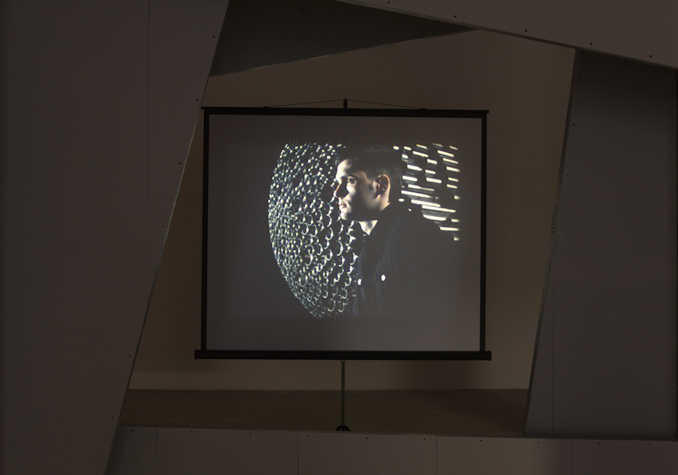 projection screen in a gallery space. on the screen there is a person stood infront of a abstract metal circle