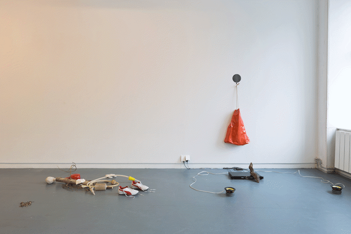 sculptural assemblage on the floor and wall of a gallery. There are trainers, sticks, plastic tubing, a hand-like object, an orange drawstring bag.