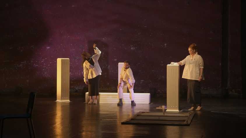 three people on stage in tramway 1 interacting with different blocks/plinths that are on the stage, one person is sat and the other two stand