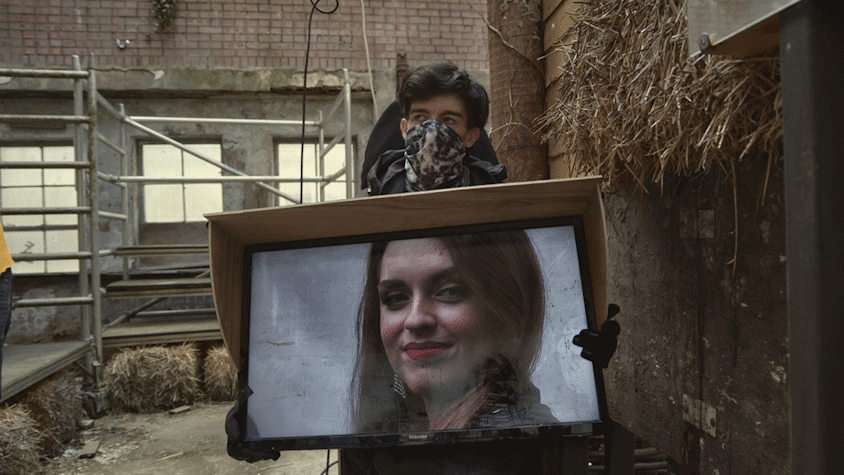 a light skinned person with dark hair and a facemask holding a tv screen with another light skinned, dark haired person with red lipstick smiling out from it