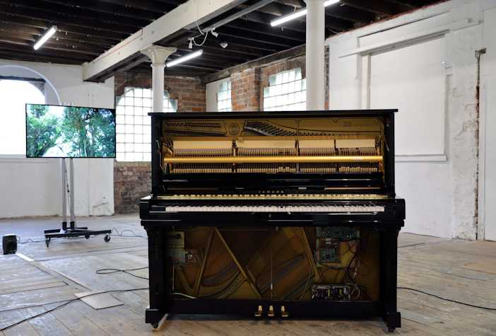 An installation of Yuko Mohri's work in the Pipe Factory, Glasgow. There is a piano and tv screen on a stand. The screen shows an image of a forest, the piano is wired up to speakers.