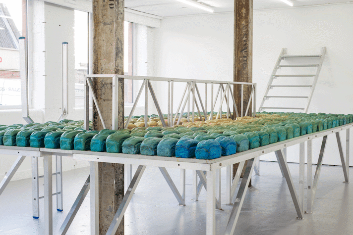 White room with metallic table in the middle, with two vertical cement beams across it. The table is filled with loafs of bread in blue and green tones.