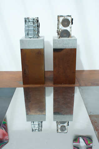 two metal sculptures on plinths next to each other. on the floor there is a mirror reflecting the sculptures.