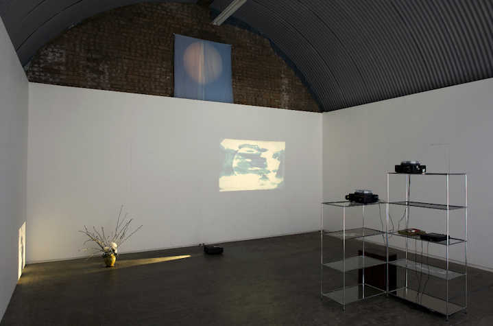 white wall in a brick space with a metal corrugated roof. there is a vase on the floor illuminated by a beam of yellow light. a projection on the wall of a red circle on a yellow and green background and two metal and glass shelves.