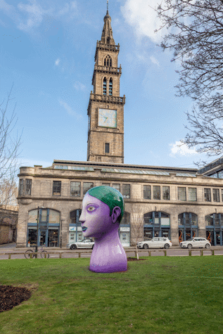 Profile right view of a big, tree-sized bust of a woman with purple skin, green eyes, thin eyebrows and green hair and lips. It is positioned in a park.