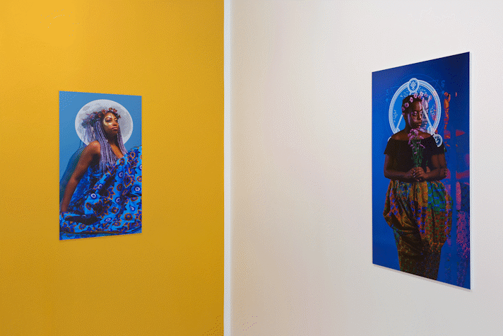 A series of portrait photographs of people of colour on bright blue backgrounds with abstract shapes around their heads, hang on a gallery wall