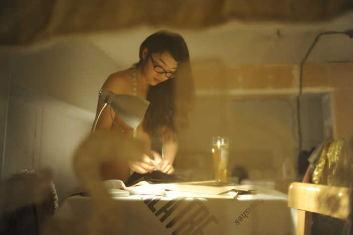 Nude person working at a dimly lit table with papers, under a desk lamp, with a drink nearby. They are wearing glasses, a measuring tape around their neck and focused on the task