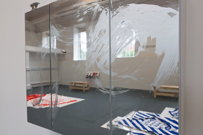 a wall mounted mirrored cabinet has a swirl of paint smeared across it and a tiny metal boat stuck to the surface