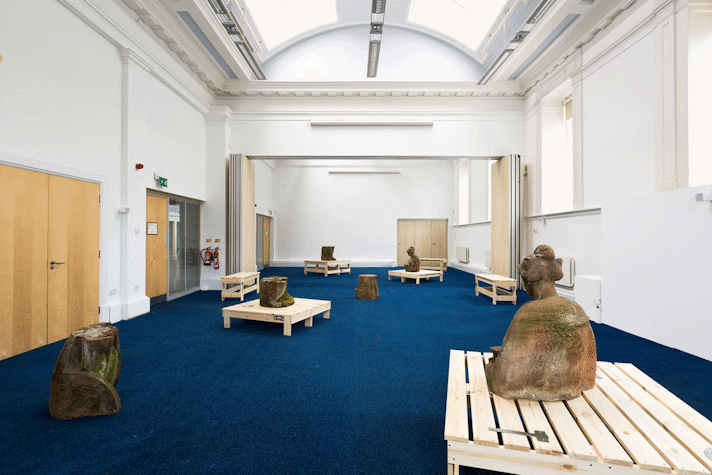In a large room with period feature ceiling and blue carpet, a series of wooden structures and sections of red sandstone sculptures are spaced out