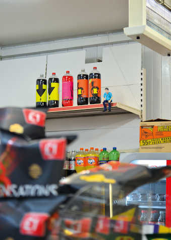 In a department store, there are drinks displayed. In the top shelf there are several colourful bottles with labels. Beside it, a small plastic figurine of a white man with blue t-shirt and jeans.