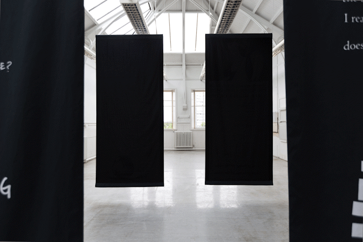 four portrait orientation large black rectangles of fabric hang from the rafters of a large school hall-esque room