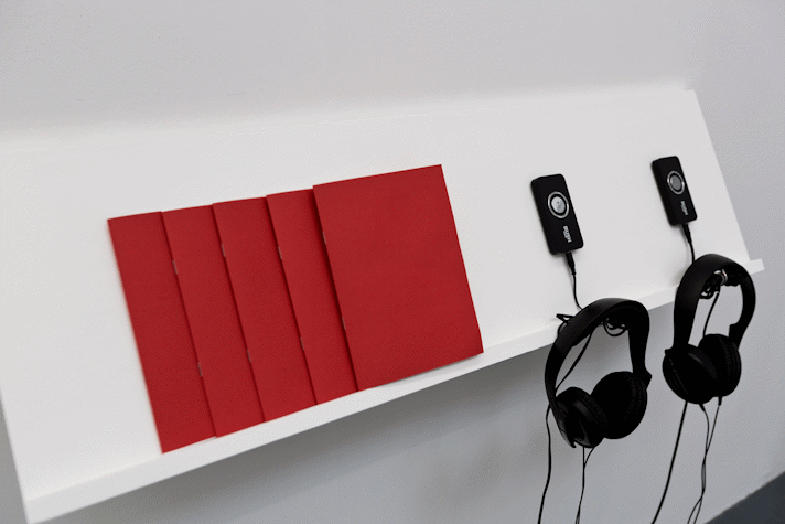 5 red covered A5 publications sit on a while shelf. Two sets of black headphones attached to mp3 players are also hanging from the shelf.