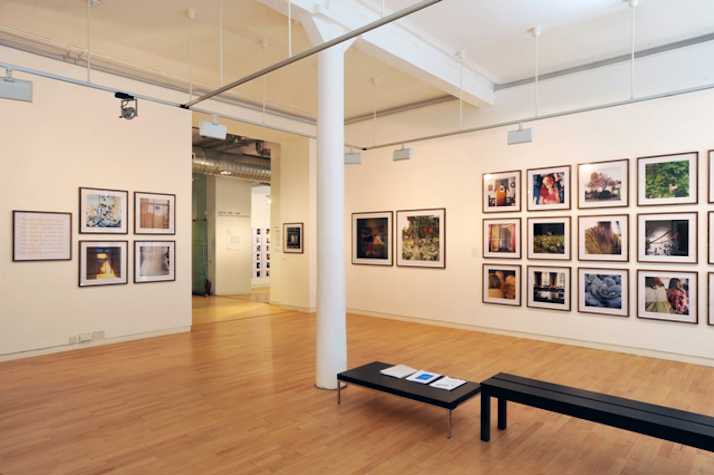 installation view with various framed photographs on the walls, a black bench and a low black table are in the centre of the space.