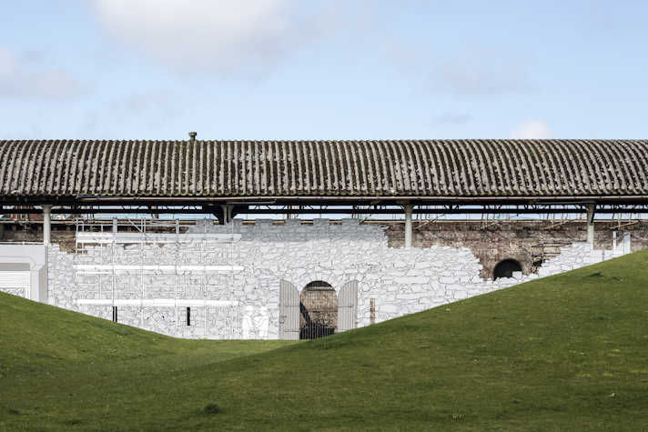 There is a blue sky with a few clouds. There is an old building and in front of it there is a black and white mural that resembles a stone wall. From left to right there is some scaffolding, a oval door and more empty stone wall. The artwork has some green grass in front of it.