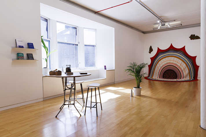 An airy white room with large bay windows. In the centre of the room is a table with two stools. At the rear wall, a large fabric print is displayed featuring circles.