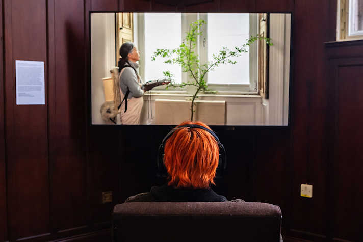The back of a person sitting on a chair watching a film on a screen, in a dark panelled room. The still on screen is a woman standing in a well lit room with a object fixed to her arm, standing in front of a tree.