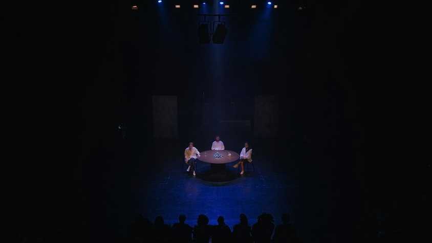 a dark theatre space. on stage there are three people sat at a roundtable.