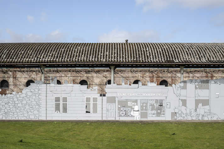 There is a blue sky with a few clouds. There is an old building and in front of it there is a black and white mural that resembles a street that depicts from left to right: a house, a TV repair and hardware store with someone looking at the window display and a building in ruins. The artwork has some green grass in front of it.