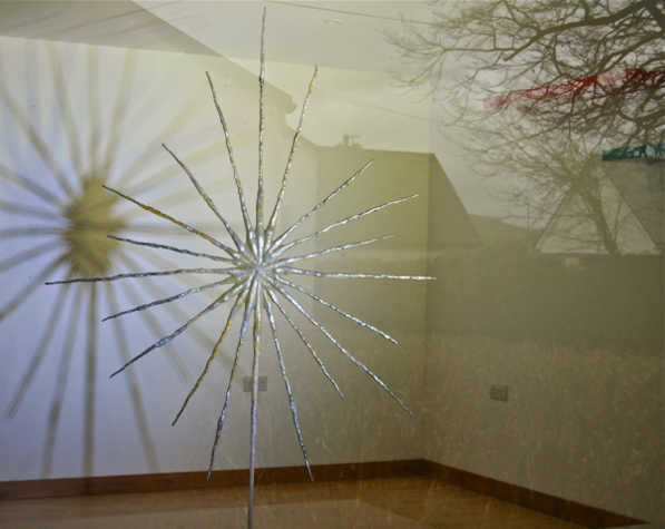 A suspended sculptural work in a star formation, casting a shadow on a white wall. The reflection of a tree and some rooftops is also visible.